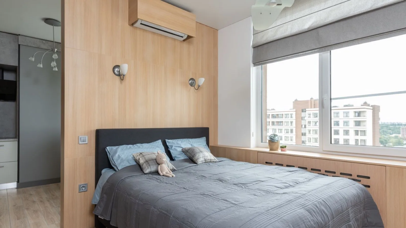 Wall-mounted 115V ductless mini split head installed high on a wood-panelled bedroom wall above the bed, with a wall outlet visible below — the kind of small room a 9,000 BTU plug-in DIY system suits