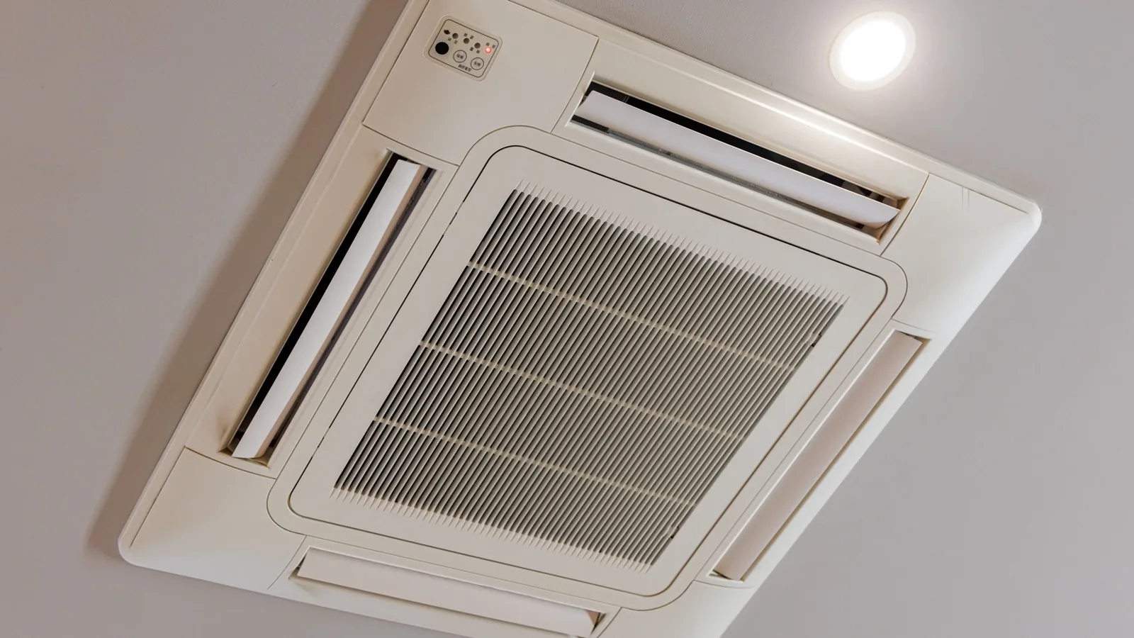 Recessed four-way ceiling cassette mini split indoor unit mounted flush in a white ceiling, its slim decorative grille and motorized louvers seen from below beside a recessed downlight