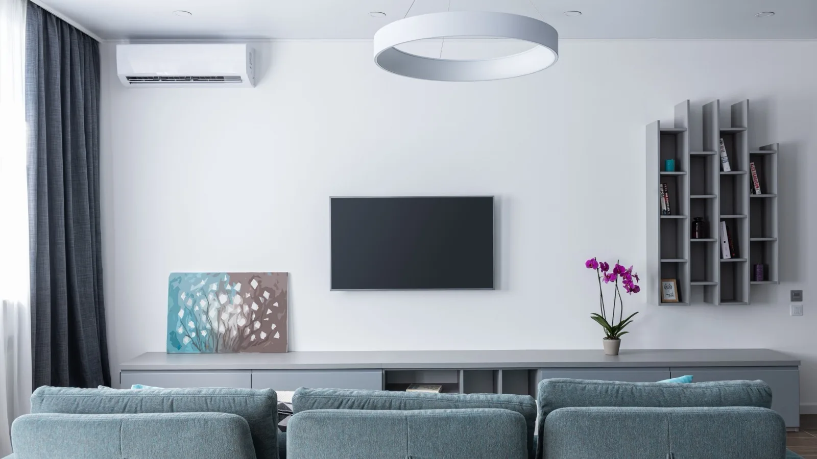 White wall-mounted ductless mini split high on the wall of a bright living room above a TV, media console and teal sofas, the kind of 500 sq ft space a 12,000 BTU DIY mini split is sized for
